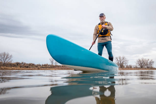 solo senior male che pagaia uno stand up paddleboard su un lago all'inizio della primavera - senior adult surfing aging process sport foto e immagini stock