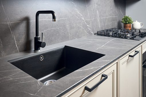 Kitchen sink area with black square matte sink tap in contemporary style. Matte black and stoneware kitchen design. Black ceramic sink with gas hob and oven, in background.