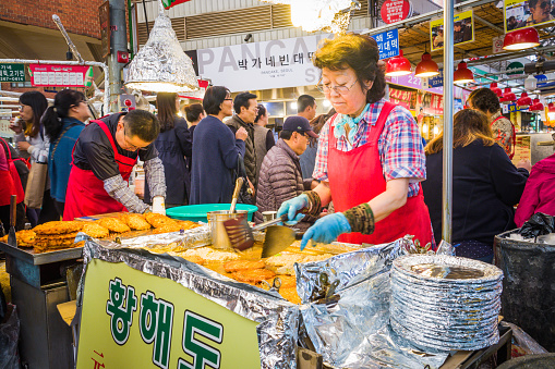 Female chef preparing traditional street food at a busy market stall in downtown Seoul, South Korea.