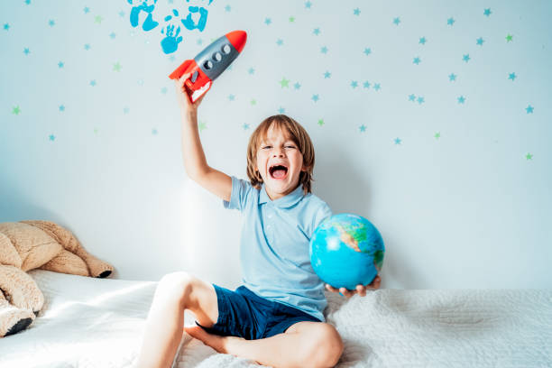 istock Emotional boy playing with space rocket and planet Earth models on bed with background of stars and family hand prints on the wall. Concept of education, future, global travel and transportation 1481204720