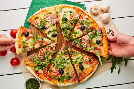 The human hand takes freshly baked pizza with ham, rukkola, sauce pesto and olives served on wooden background with tomatoes, sauces and herbs. Food delivery concept