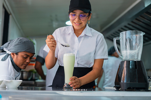 Average age 25-year-old Latino female kitchen assistant in a restaurant dressed in a kitchen uniform is preparing delicious lemonade for customers