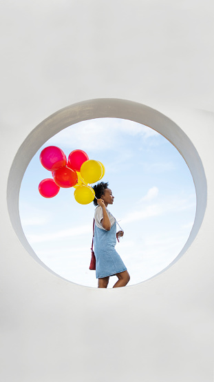 Young woman standing outside framed in a circle in a wall and holding on to a bunch of red and yellow balloons