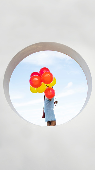 Young woman framed in a circle in a wall outdoors and holding on to a bunch of red and yellow balloons