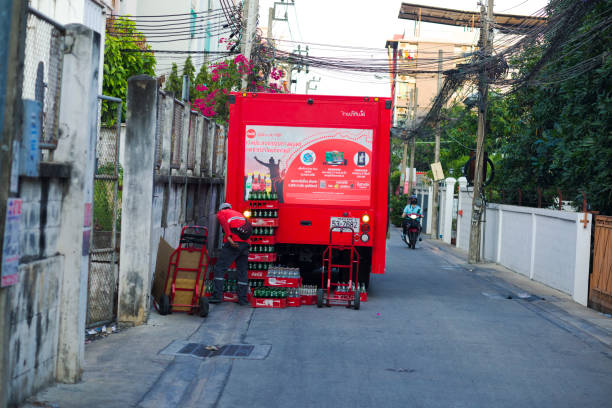 Coca Cola truck and delivering staff is standing in street in residential district in Bangkok Chatuchak near CRU university. Truck is delivering boxes with bottles and re-collecting empty bottles. Truck is standing in front of larger condominium