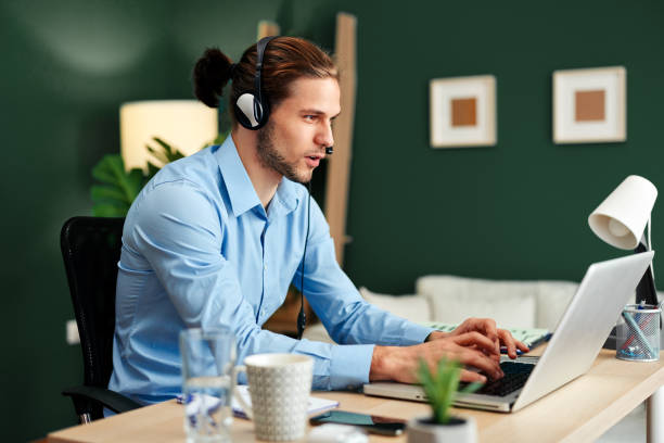 Un joven con auriculares y una computadora portátil tiene una llamada de conferencia en la oficina - foto de stock