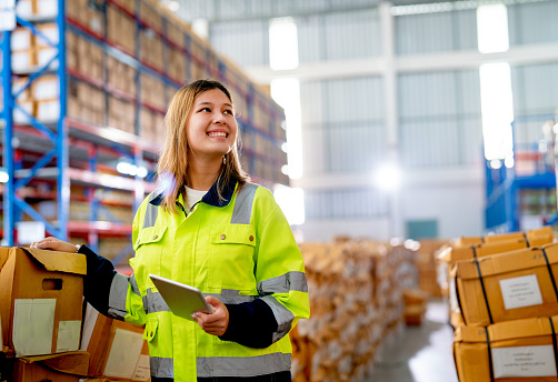 Beautiful young warehouse worker woman hold tablet and smiling also look to her left side in workplace area.