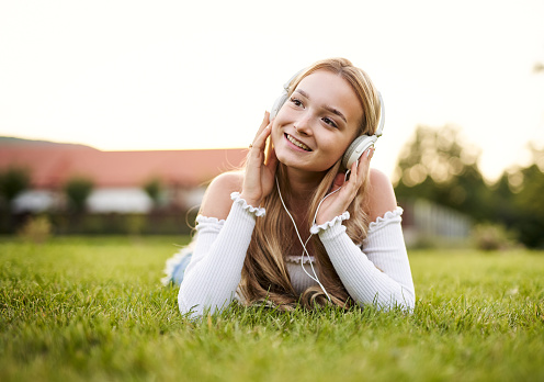 Teenager girl enjoying life and lying on front in the grass in a public park, while listening to music with headphones on her head with the sunset in the background on a sunny day