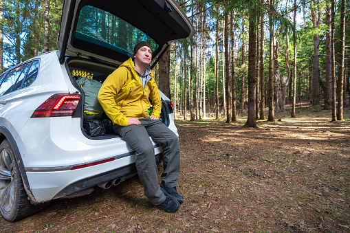 An adult man sitting on the car’s trunk and enjoys the view of solitude camping site in the woods.