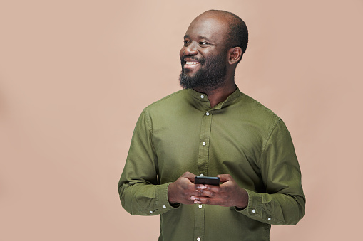 Portrait of happy African American man using smartphone and smiling while standing on brown background