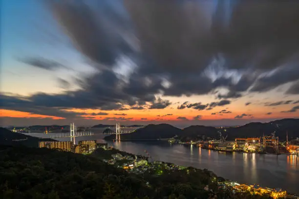 Long exposure sunset photography of Japanese Megami cable-stayed bridge in Nagasaki bay taken from Mt Nabekanmuri's observation deck surrounded by mountain sides and illuminated shipyards.