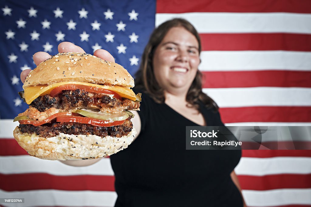 Gordo jovem smilingly oferece hambúrguer de contra Bandeira dos EUA - Foto de stock de Bandeira Norte-Americana royalty-free