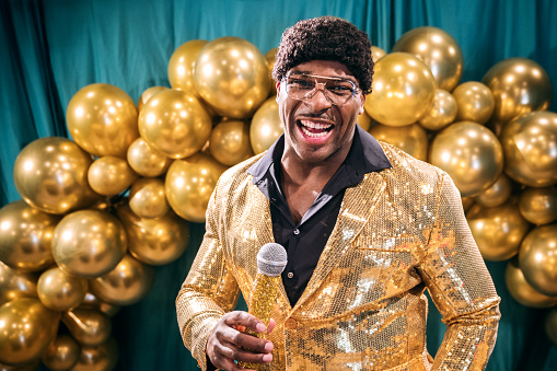 A fun depiction of a competitive TV game show, stylized in late 1970's or early 1980's fashion.  The host, an African American man in a stunning gold blazer, talks to the crowd and the television.