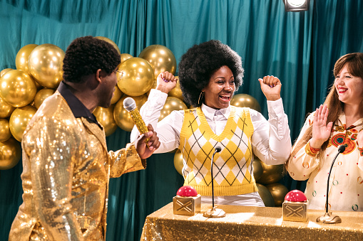 A fun depiction of a competitive TV game show, stylized in late 1970's or early 1980's fashion.  The host, an African American man in a stunning gold blazer, asks the contestants quiz trivia questions to see who will win the grand prize!