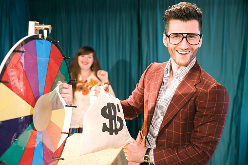 A fun depiction of a competitive TV game show, stylized in late 1970's or early 1980's fashion.   Contestants spin a colored wheel to win cash prizes.  A Caucasian man holds up a money bag he has won.