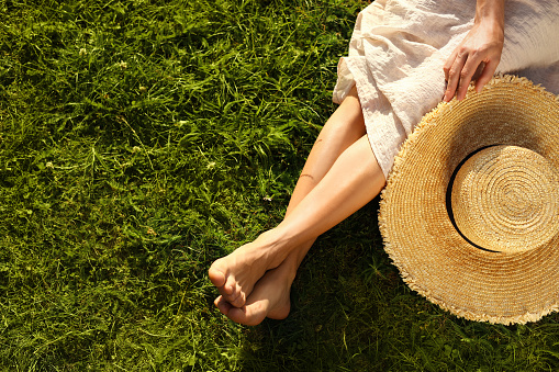 Woman with hat sitting barefoot on green grass outdoors, top view. Space for text