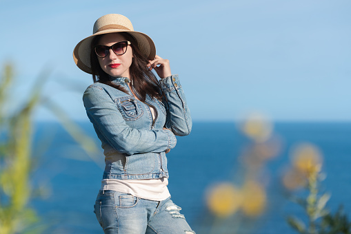 Woman in sunglasses, straw hat, denim jacket and jeans pose beautifully against backdrop of blue sky and ocean. 40 year old hipster woman on vacation. Frame of green grass around edges of photo. Part of a series