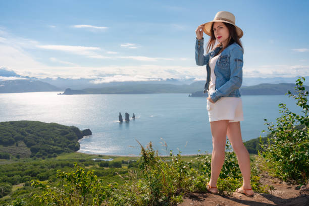 Hipster female standing on high mountain against backdrop of panoramic ocean on sunny summer day stock photo