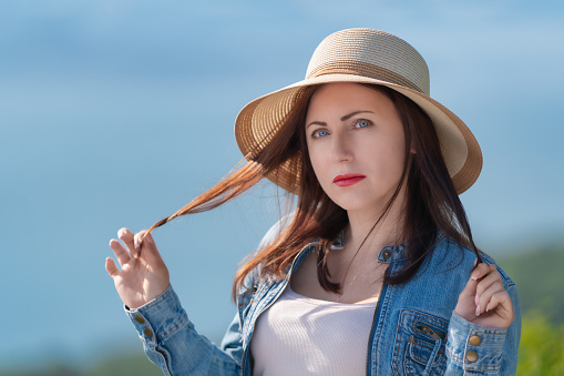 Mysterious adult woman with gray eyes looking at camera. Hipster dressed in straw hat, denim jacket and light T-shirt. Female tourist standing on blurred natural background of sea and forest. Part of a series