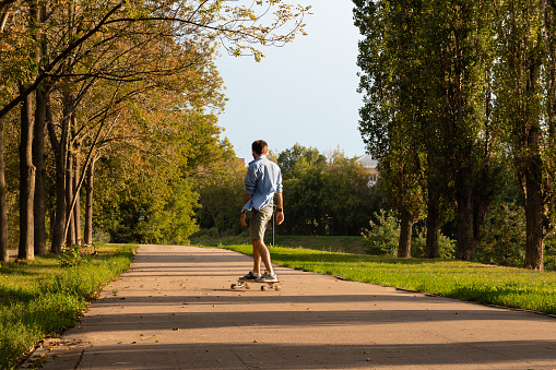 Back view of unrecognizable young guy riding on longboard along empty asphalt road between green trees