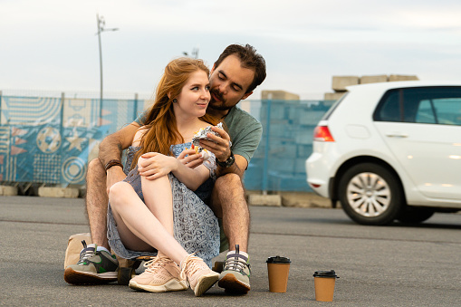 Young couple sitting on skateboard together and eating takeaway food. Woman looking away with smiling and man looking at the woman.