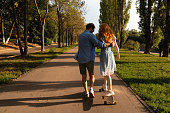 Young man helping girlfriend to ride longboard on sunny alley against green trees