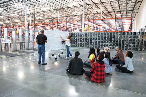 A mature African American man and a mature white woman present to a diverse group of employees in a fulfillment center.