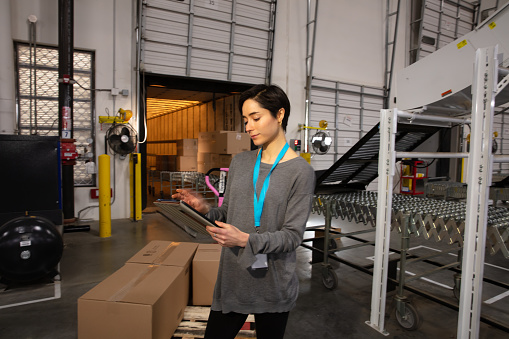 A Hispanic woman with a digital tablet standing next to a dolly with cardboard boxes in a fulfillment center.