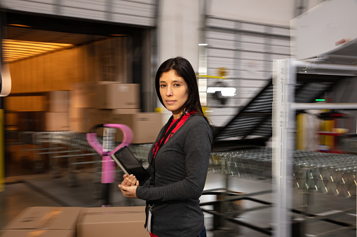 A Hispanic woman with a digital tablet standing next to cardboard boxes in a fulfillment center.