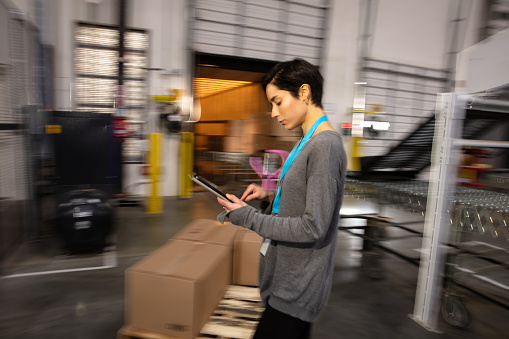 A Hispanic woman with a digital tablet standing next to a dolly with cardboard boxes in a fulfillment center.