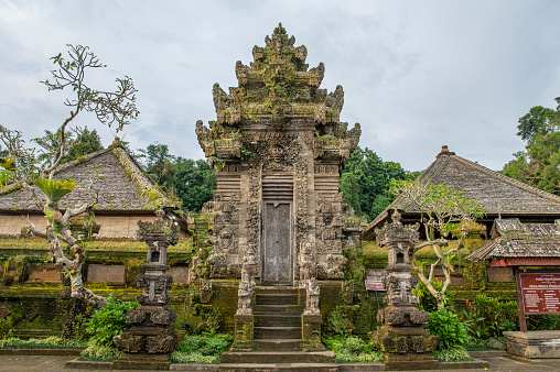 The dark stone, adorned with intricate carvings, frames the view beyond. A concrete pathway leads through the center of the gateway, inviting visitors to explore. In the background, misty mountains partially shrouded in clouds create a serene and mystical atmosphere. The scene captures the essence of Balinese spirituality and natural beauty.