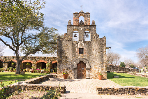 Mission Espada bathed in sunlight, San Antonio, Texas. Stone front of mission with bells. Blue sky with clouds, mature tree, and stone walkway of the Mission
