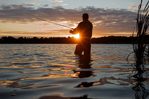 Fisherman wearing a chest wader standing in the lake fishing at sunset with lens flare defect.