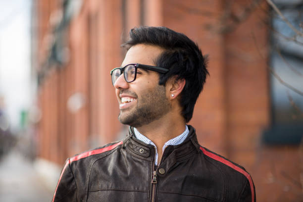 Smiling Indian ethnicity man with an earring, eyeglasses and wearing a leather jacket over a dress shirt is looking to his right out at the road as he is standing on the sidewalk of a city near tall brick buildings in the spring.