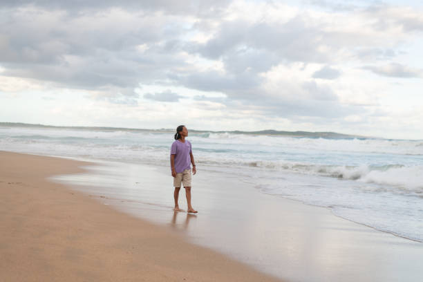 Wide shot of a middle age male walking by the ocean, barefoot and by himself.  Cloudy background and waves in the ocean.
