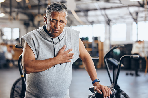 Sports, gym and senior man with pain in chest, medical emergency during workout at fitness studio. Health, wellness and inflammation, old man with hand on heart cramps while training on exercise bike