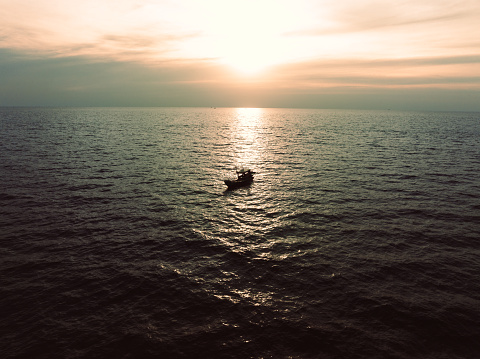 Thai fisherman sitting in thai fishing longtail boat starting to fish during sunset in the Andaman Sea close to Khao Lac Island under moody colorful dusk skyscape. Drone Point of View. Moody sunset over the Andaman Sea. Khao Lak Island, Andaman Sea, Phuket, Thailand, Southeast Asia.