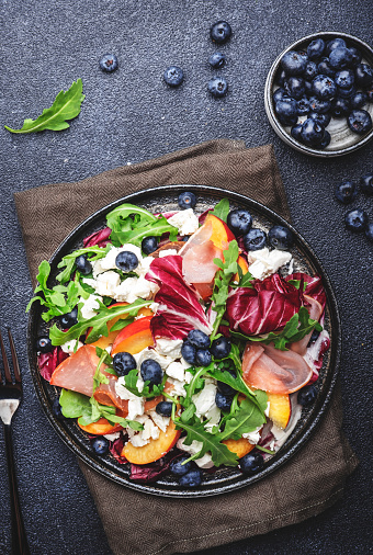 Delicious salad  with arugula, radicchio, peaches, prosciutto, feta cheese and blueberries. Black table background, top view