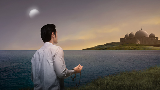 Muslim man standing while raised hands holding prayer beads and praying with mosque background