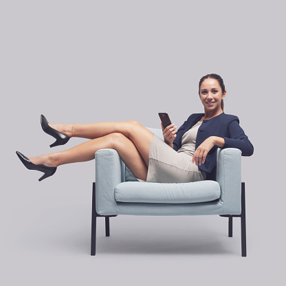 Confident young businesswoman sitting on a chair with feet up and holding a smartphone, she is smiling at camera