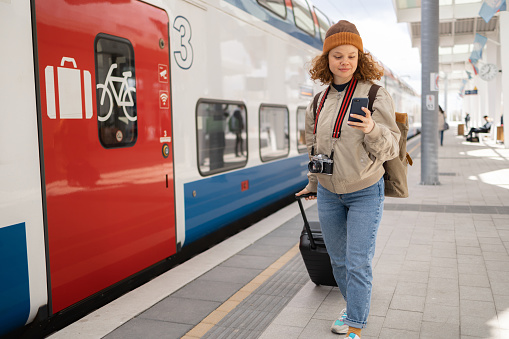 Young female backpacker using smart phone while pulling her luggage on a train station.