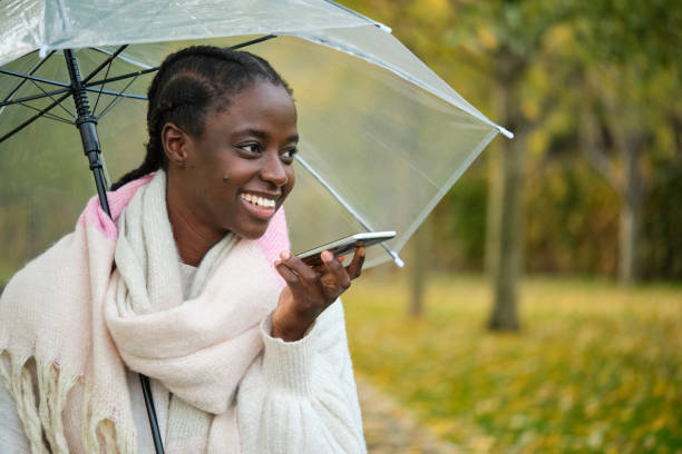 donna africana con un ombrello che sorride e registra un messaggio audio in autunno. - spanish culture audio foto e immagini stock