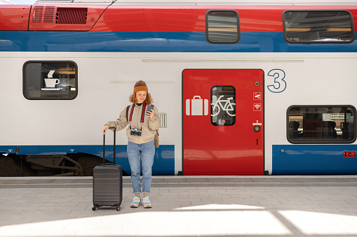 Smiling female tourist text messaging on mobile phone while standing with her luggage on a train station.