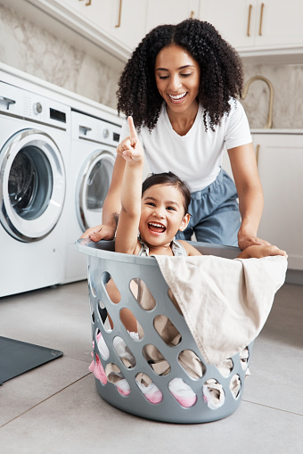 Mom, laundry and girl kid in basket by washing machine for cleaning, bonding and comic time in house. Crazy fun, mother and daughter with happiness, love and care in family home with smile on floor