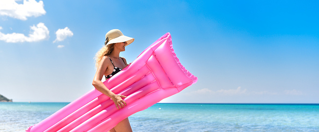 Happy woman and wearing beach hat with pink mattress having summer fun during travel holidays.\nHappy woman with inflatable pink mattress on the beach.
