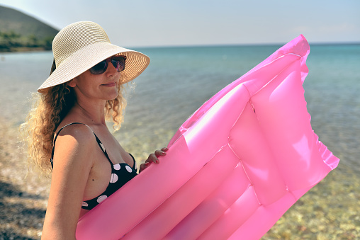 Happy woman and wearing beach hat with pink mattress having summer fun during travel holidays.
Happy woman with inflatable pink mattress on the beach.