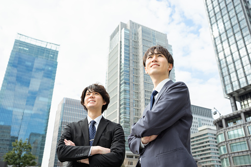 Two men in a suit in their 20s and 30s who look far away in a city surrounded by skyscrapers and make their arms assembled with a smile.