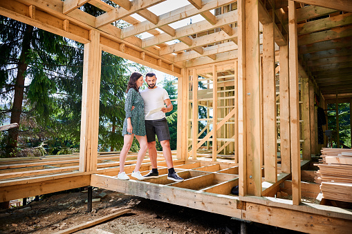 Man and woman examining their future wooden frame dwelling nestled near the forest. Youthful couple at construction site in early morning. Concept of contemporary ecological construction.
