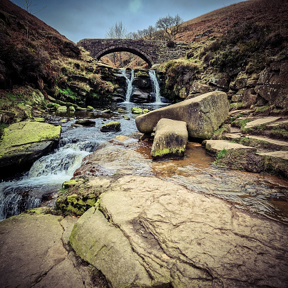 Three Shire Heads, where water cascades over mossy rocks and a packhorse bridge arches over the stream. Lush, rolling hills of the Peak District embrace this enchanting meeting point