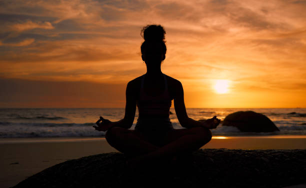Sunset, beach and silhouette of a woman in a lotus pose while doing a yoga exercise by the sea. Peace, zen and shadow of a calm female doing meditation or pilates workout outdoor at dusk by the ocean stock photo